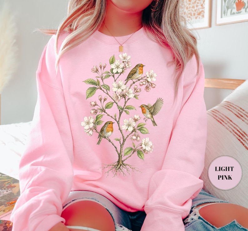 May include: Light pink sweatshirt featuring a detailed illustration of a flowering tree with three birds perched on its branches. The birds have orange breasts and brown wings. The tree has white blossoms and green leaves.