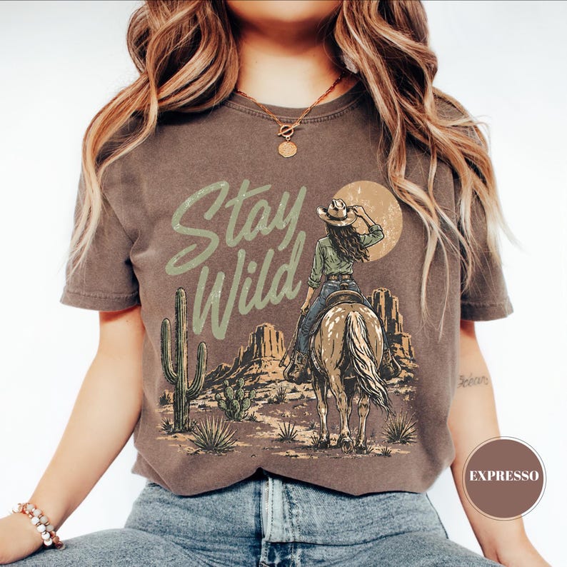 May include: Brown t-shirt with a desert graphic. The design shows a cowgirl on horseback, cacti, and the words "Stay Wild" in light green. The shirt has a vintage, worn appearance.