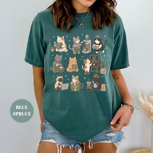 May include: Blue spruce t-shirt featuring a whimsical design of cats engaged in coffee-related activities. The cats are depicted reading, brewing coffee, and enjoying their beverages. The shirt includes the text "Coffee First, Everything Else".