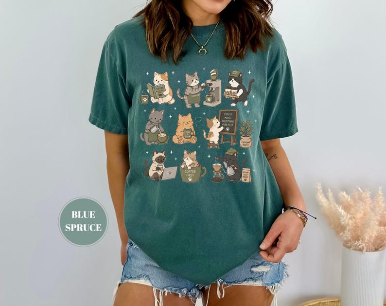 May include: Blue spruce t-shirt featuring a whimsical design of cats engaged in coffee-related activities. The cats are depicted reading, brewing coffee, and enjoying their beverages. The shirt includes the text "Coffee First, Everything Else".