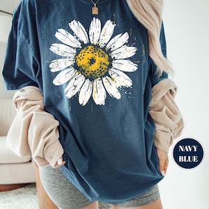 Puede incluir: Camiseta azul marino con una gran margarita blanca y centro amarillo. El dise&ntilde;o de la margarita tiene un aspecto desgastado y vintage. La camiseta tiene cuello redondo y mangas cortas. Las palabras "NAVY BLUE" est&aacute;n en un c&iacute;rculo azul marino.