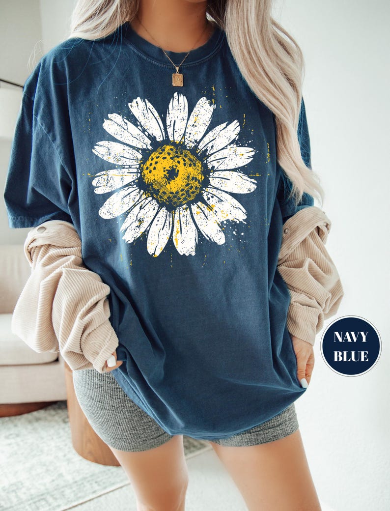 Puede incluir: Camiseta azul marino con una gran margarita blanca y centro amarillo. El dise&ntilde;o de la margarita tiene un aspecto desgastado y vintage. La camiseta tiene cuello redondo y mangas cortas. Las palabras "NAVY BLUE" est&aacute;n en un c&iacute;rculo azul marino.