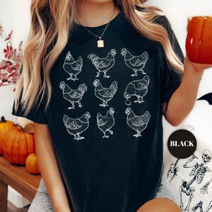 May include: Black t-shirt featuring a grid of nine white chicken illustrations. The shirt has a crew neck and short sleeves. A round sticker on the shirt says "BLACK". The shirt is worn by a person.