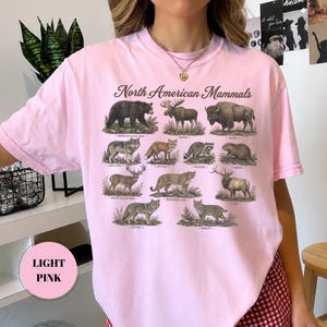 May include: A light pink t-shirt featuring the text North American Mammals above a grid of illustrations of various animals including a black bear, moose, bison, gray wolf, red fox, raccoon, beaver, white-tailed deer, mountain lion, elk, coyote, and bobcat.