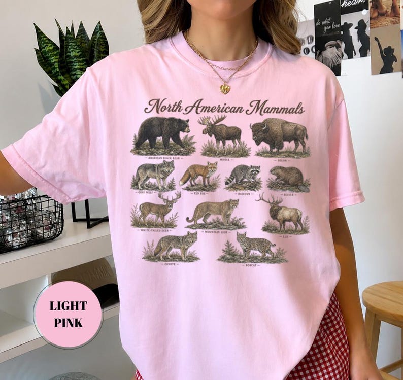 May include: A light pink t-shirt featuring the text North American Mammals above a grid of illustrations of various animals including a black bear, moose, bison, gray wolf, red fox, raccoon, beaver, white-tailed deer, mountain lion, elk, coyote, and bobcat.