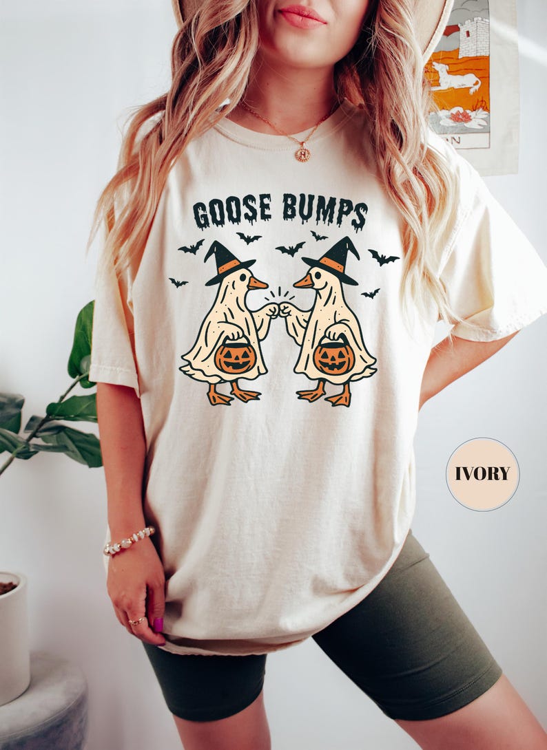 May include: Ivory-colored t-shirt with the text "GOOSE BUMPS" above a graphic of two ducks in witch hats bumping fists, each holding a jack-o'-lantern. Black bats are scattered around the ducks. A fun Halloween-themed tee.