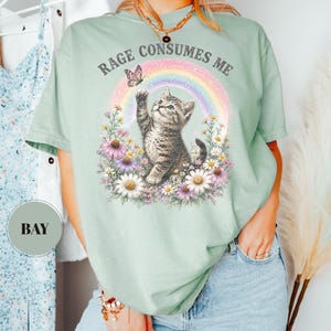 May include: Sage green t-shirt featuring a kitten reaching for a butterfly in front of a rainbow and flowers. The text "RAGE CONSUMES ME" is arched above the design. The shirt is a relaxed fit.