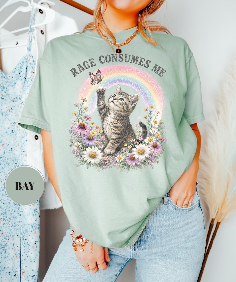 May include: Sage green t-shirt featuring a kitten reaching for a butterfly in front of a rainbow and flowers. The text "RAGE CONSUMES ME" is arched above the design. The shirt is a relaxed fit.