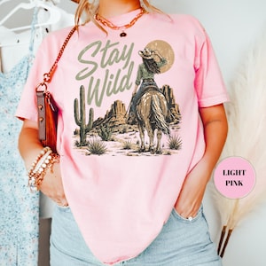 May include: Pale pink t-shirt featuring a desert scene with a person on horseback and the words "Stay Wild". The shirt has a loose fit and is made of a soft material.