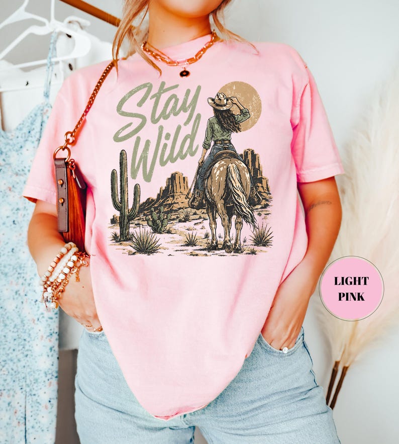 May include: Pale pink t-shirt featuring a desert scene with a person on horseback and the words "Stay Wild". The shirt has a loose fit and is made of a soft material.