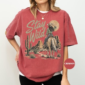 May include: Crimson red t-shirt with a desert graphic. The design shows a cowgirl on horseback, a cactus, and the words "Stay Wild" in a distressed font. The shirt has short sleeves and a crew neck.