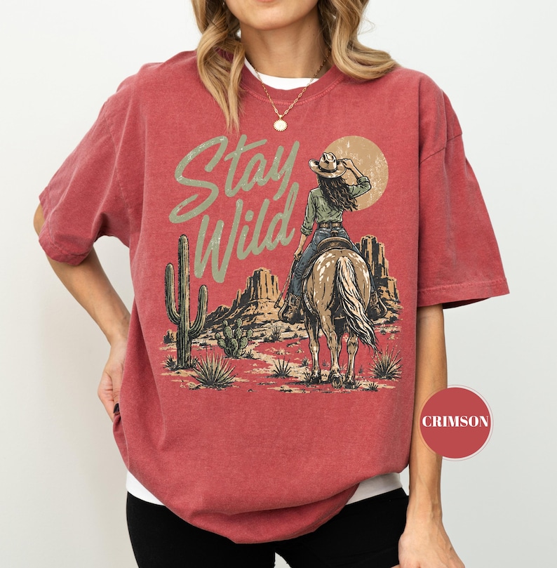 May include: Crimson red t-shirt with a desert graphic. The design shows a cowgirl on horseback, a cactus, and the words "Stay Wild" in a distressed font. The shirt has short sleeves and a crew neck.