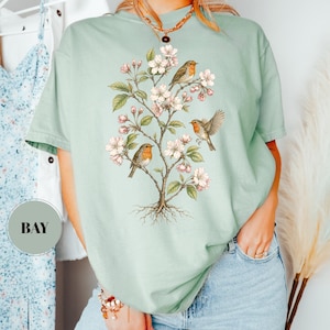 May include: A light green t-shirt featuring a detailed illustration of a flowering tree with three robins perched on its branches. The tree has white blossoms and green leaves. The shirt is a comfortable, casual style.