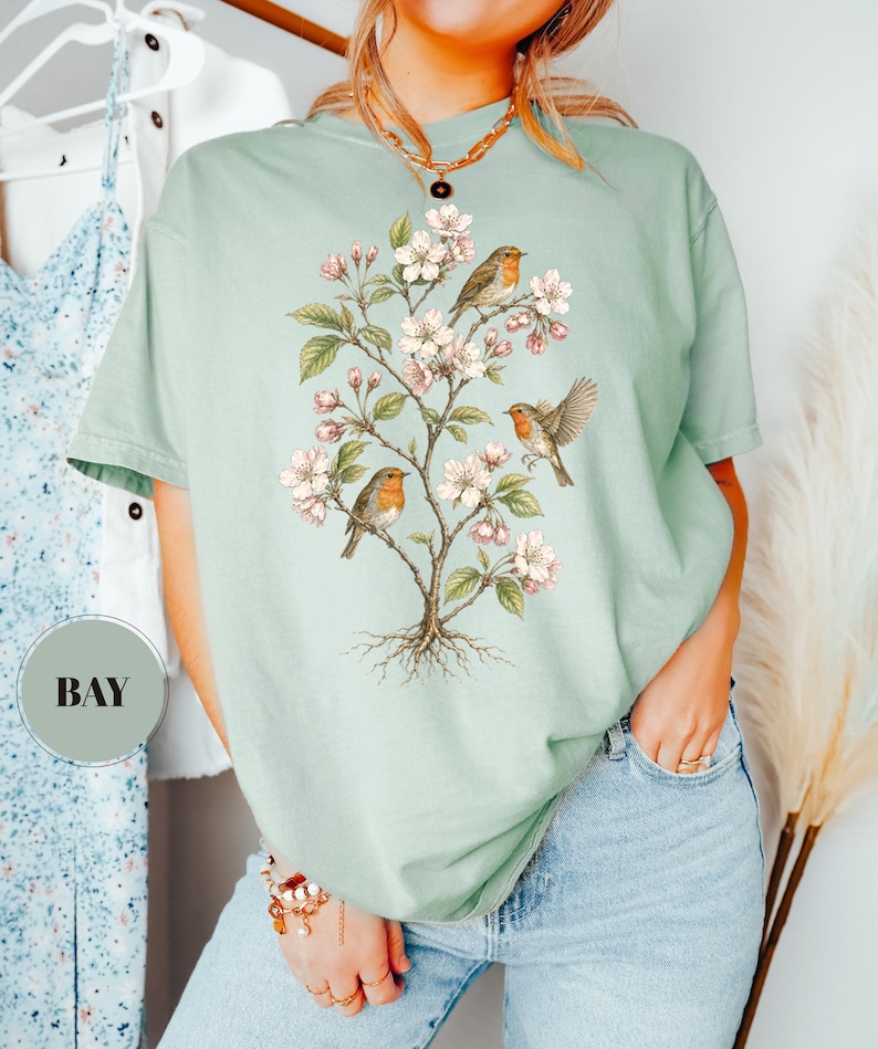 May include: A light green t-shirt featuring a detailed illustration of a flowering tree with three robins perched on its branches. The tree has white blossoms and green leaves. The shirt is a comfortable, casual style.