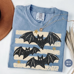 May include: A blue short-sleeved t-shirt with a graphic print of three black bats and gold stars over horizontal cream-colored stripes. The inner neck label reads Comfort Colors, 100% cotton, S P CH. A gold-toned necklace rests on the fabric.