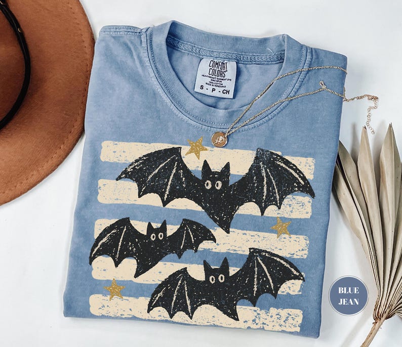 May include: A blue short-sleeved t-shirt with a graphic print of three black bats and gold stars over horizontal cream-colored stripes. The inner neck label reads Comfort Colors, 100% cotton, S P CH. A gold-toned necklace rests on the fabric.