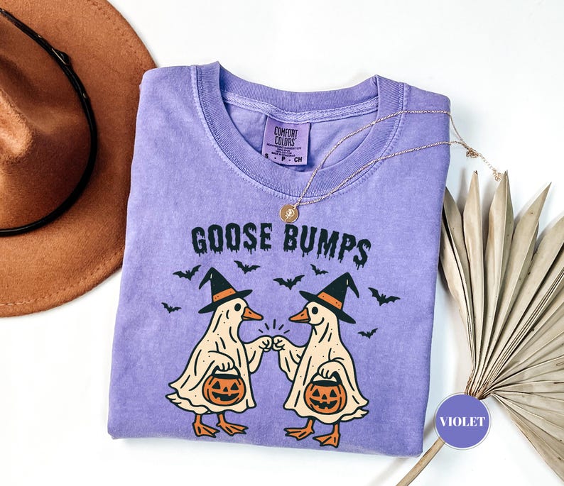 May include: Violet-colored t-shirt with the words "GOOSE BUMPS" and a graphic of two cartoon geese wearing witch hats and holding pumpkin buckets. The shirt has a crew neck and short sleeves. A gold necklace and a brown hat are also visible.
