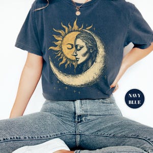 May include: Navy blue t-shirt featuring a sun and moon graphic. The design depicts a sun with a face and a woman's face within a crescent moon. The shirt is a dark blue color.