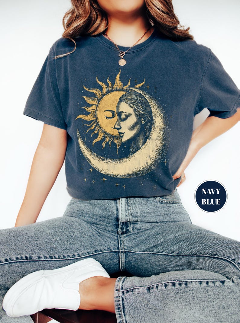 May include: Navy blue t-shirt featuring a sun and moon graphic. The design depicts a sun with a face and a woman's face within a crescent moon. The shirt is a dark blue color.