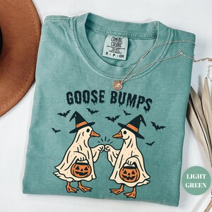 May include: Light green t-shirt with the text "GOOSE BUMPS" and a graphic of two cartoon geese wearing witch hats and holding jack-o'-lanterns. The geese are bumping fists. Bats are flying around the geese. A gold necklace is on the shirt.