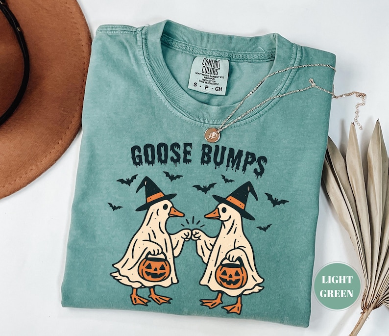 May include: Light green t-shirt with the text "GOOSE BUMPS" and a graphic of two cartoon geese wearing witch hats and holding jack-o'-lanterns. The geese are bumping fists. Bats are flying around the geese. A gold necklace is on the shirt.