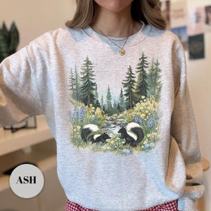 May include: Ash-colored sweatshirt featuring a nature scene with two skunks, a stream, and trees. The design includes blue and yellow wildflowers. The word "ASH" is printed on a circular label at the bottom.