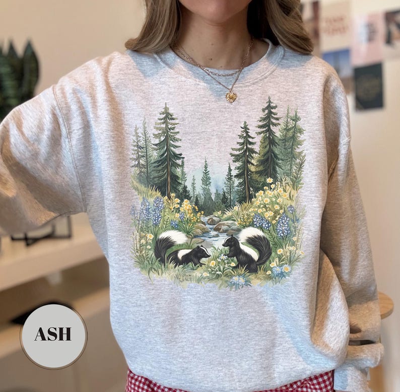 May include: Ash-colored sweatshirt featuring a nature scene with two skunks, a stream, and trees. The design includes blue and yellow wildflowers. The word "ASH" is printed on a circular label at the bottom.