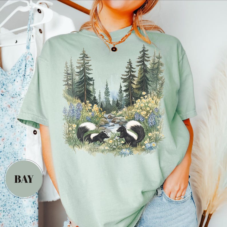 May include: A light green t-shirt featuring a nature scene with two skunks, a stream, and a forest. The skunks are black and white, and the forest has tall green trees. The shirt has the word "BAY" printed on it.