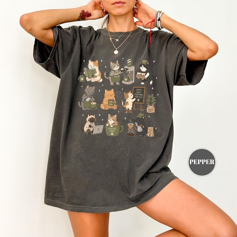 May include: Dark grey t-shirt featuring a whimsical illustration of cats engaged in various coffee-related activities. The cats are depicted reading, brewing coffee, and working on a laptop. The shirt has a relaxed fit.