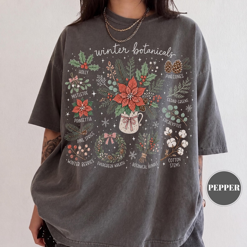May include: A charcoal gray short-sleeve t-shirt featuring a graphic print titled winter botanicals. The design includes illustrations of holly, mistletoe, poinsettia, pine sprigs, winter berries, pinecones, cedar greens, eucalyptus, cotton stems, and a wreath.