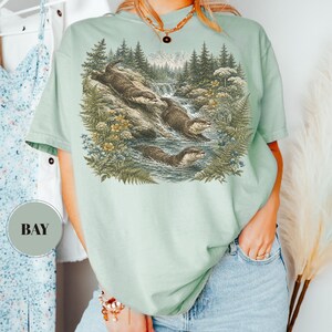Puede incluir: Una camiseta de manga corta de color verde claro con un estampado gr&aacute;fico detallado de tres nutrias nadando en un arroyo forestal con pinos, helechos y flores silvestres. La camiseta tiene cuello redondo y una etiqueta circular con el texto BAY.