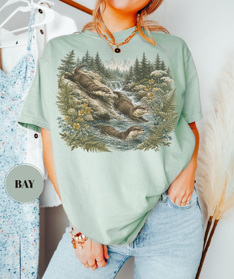 Puede incluir: Una camiseta de manga corta de color verde claro con un estampado gr&aacute;fico detallado de tres nutrias nadando en un arroyo forestal con pinos, helechos y flores silvestres. La camiseta tiene cuello redondo y una etiqueta circular con el texto BAY.