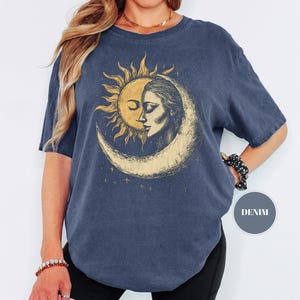May include: A denim blue t-shirt featuring a celestial design. The graphic depicts a sun and moon with a woman's face. The design is in shades of gold and cream. The shirt has short sleeves and a round neckline.