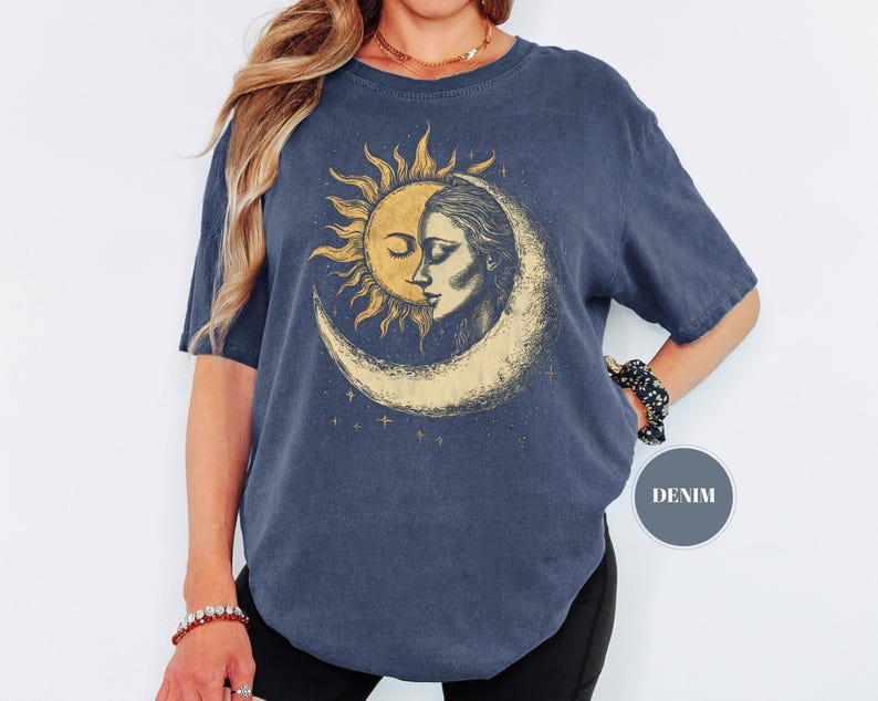 May include: A denim blue t-shirt featuring a celestial design. The graphic depicts a sun and moon with a woman's face. The design is in shades of gold and cream. The shirt has short sleeves and a round neckline.