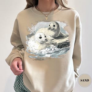 Pode incluir: Camisola cor de areia com uma ilustra&ccedil;&atilde;o vintage de duas focas numa rocha no oceano. As focas s&atilde;o brancas e cinzentas, com pelo detalhado e olhos expressivos. A camisola tem gola redonda e mangas compridas.