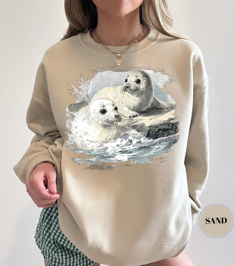 Pode incluir: Camisola cor de areia com uma ilustra&ccedil;&atilde;o vintage de duas focas numa rocha no oceano. As focas s&atilde;o brancas e cinzentas, com pelo detalhado e olhos expressivos. A camisola tem gola redonda e mangas compridas.