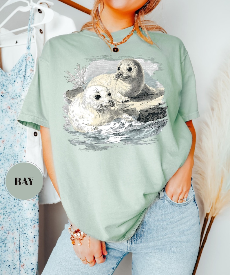 Pode incluir: T-shirt verde s&aacute;lvia com uma ilustra&ccedil;&atilde;o vintage de duas focas numa rocha no oceano. As focas s&atilde;o brancas com olhos escuros, e a obra de arte &eacute; detalhada com sombreamento e linhas. A t-shirt tem um corte descontra&iacute;do.