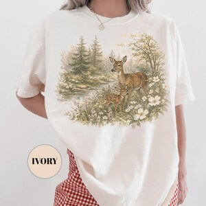 May include: Ivory-colored t-shirt featuring a detailed illustration of a deer and fawn in a forest setting. The artwork includes a river, trees, and white flowers. The shirt has short sleeves and a round neckline. The word "IVORY" is printed on a circle.