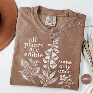以下が含まれることがあります： 植物のボタニカルイラストと「all plants are edible some only once」というテキストが白でプリントされたブラウンのTシャツ。クルーネックで、襟の内側にはComfort Colorsのブランドラベルが付いています。
