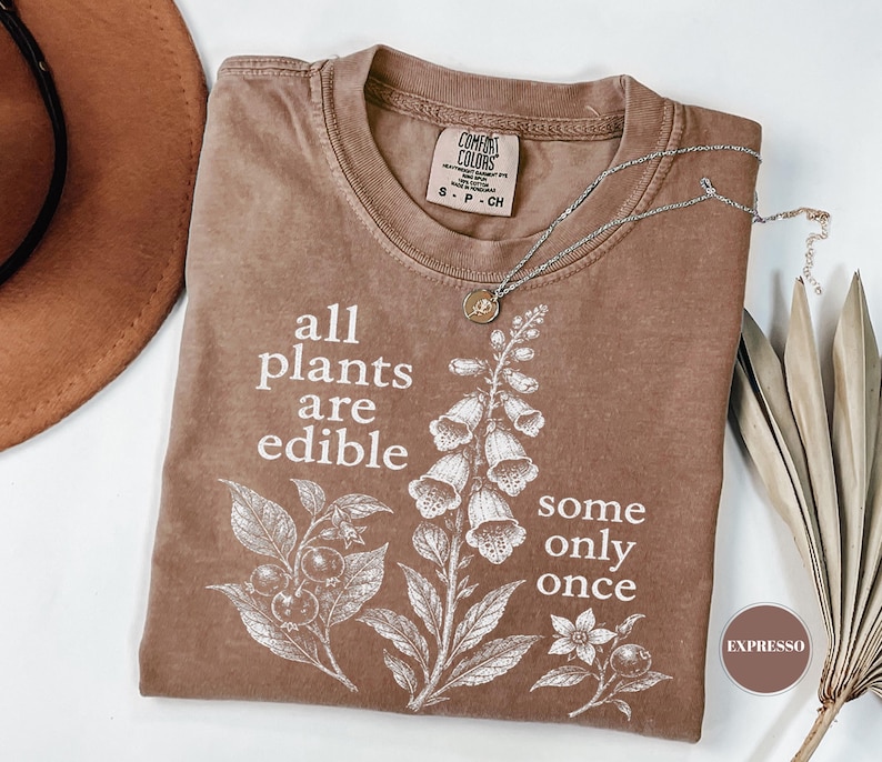 以下が含まれることがあります： 植物のボタニカルイラストと「all plants are edible some only once」というテキストが白でプリントされたブラウンのTシャツ。クルーネックで、襟の内側にはComfort Colorsのブランドラベルが付いています。