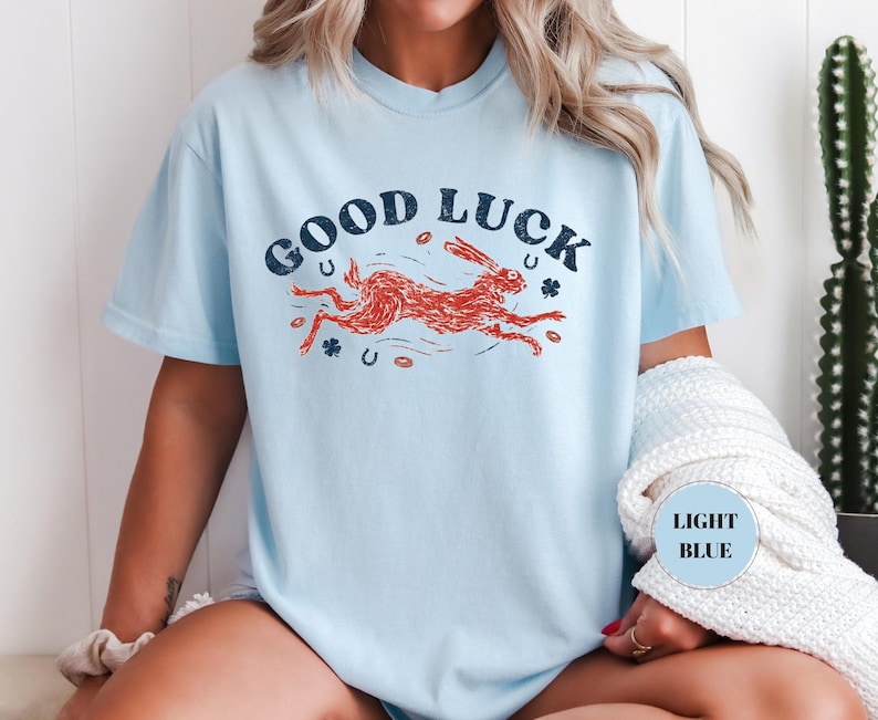K&ouml;nnte beinhalten: Hellblaues T-Shirt mit den Worten "GOOD LUCK" in marineblauem, gebogenem Text. In der Mitte befindet sich ein roter, rennender Hase mit Kleebl&auml;ttern und Hufeisen. Das Shirt hat eine hellblaue Farbe.