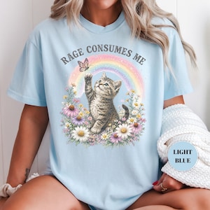 May include: Light blue t-shirt featuring a kitten reaching for a butterfly above a rainbow. The text "RAGE CONSUMES ME" arches above the design. The kitten is surrounded by colorful flowers. The shirt has a relaxed fit.