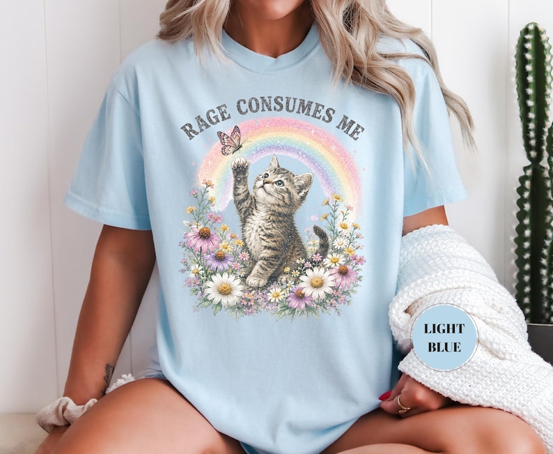 May include: Light blue t-shirt featuring a kitten reaching for a butterfly above a rainbow. The text "RAGE CONSUMES ME" arches above the design. The kitten is surrounded by colorful flowers. The shirt has a relaxed fit.