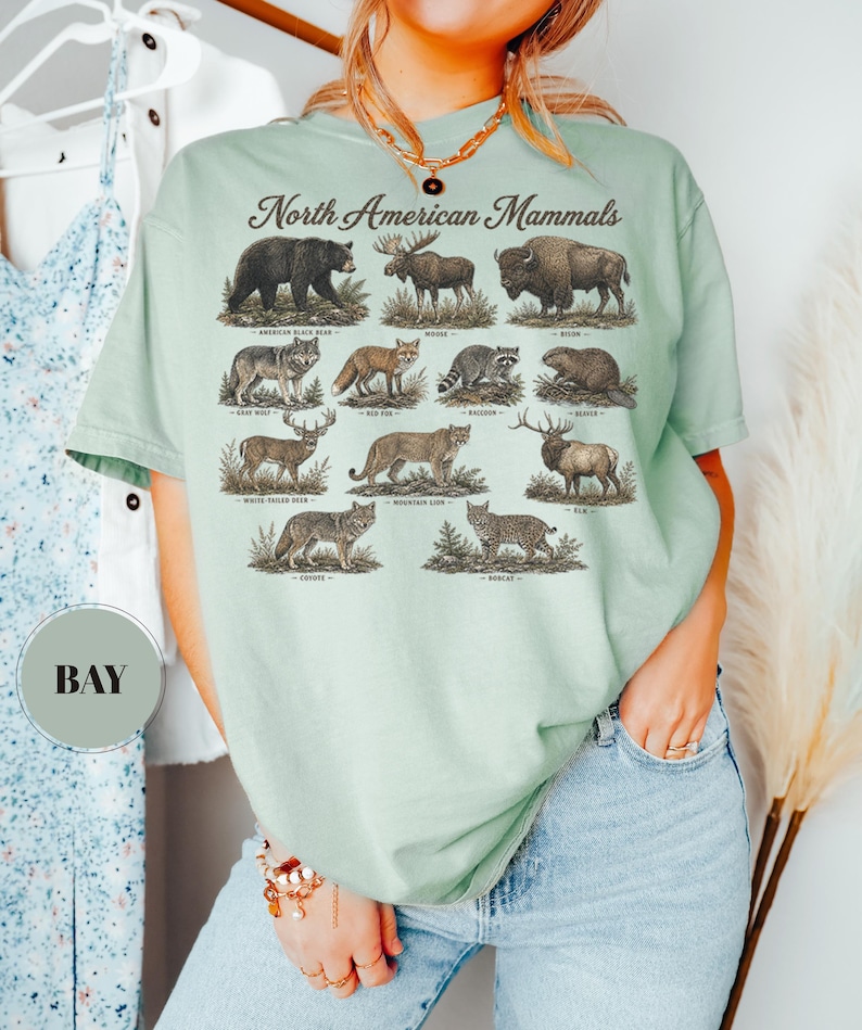 May include: A light green t-shirt featuring a graphic print titled North American Mammals. The design displays illustrations of an American black bear, moose, bison, gray wolf, red fox, raccoon, beaver, white-tailed deer, mountain lion, elk, coyote, and bobcat.