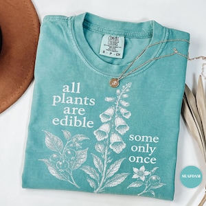 以下が含まれることがあります： シーフォームグリーンの半袖Tシャツ。植物のイラストと「all plants are edible some only once」という白いグラフィックプリントが施されています。クルーネックで、襟の内側にはComfort Colorsのラベルが付いています。