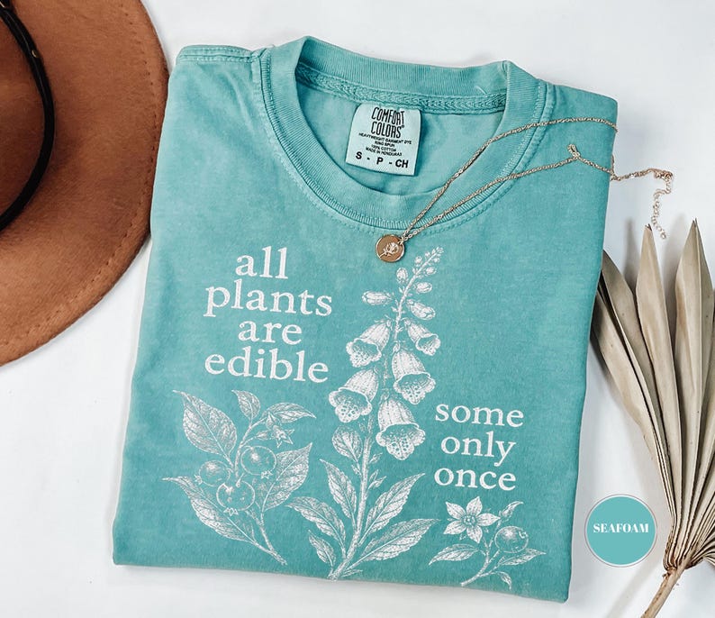 以下が含まれることがあります： シーフォームグリーンの半袖Tシャツ。植物のイラストと「all plants are edible some only once」という白いグラフィックプリントが施されています。クルーネックで、襟の内側にはComfort Colorsのラベルが付いています。