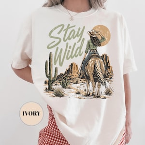 May include: Ivory t-shirt with a desert-themed graphic. It shows a cowgirl on horseback, a cactus, and the words "Stay Wild" in green. The design also includes a sun and mountains.
