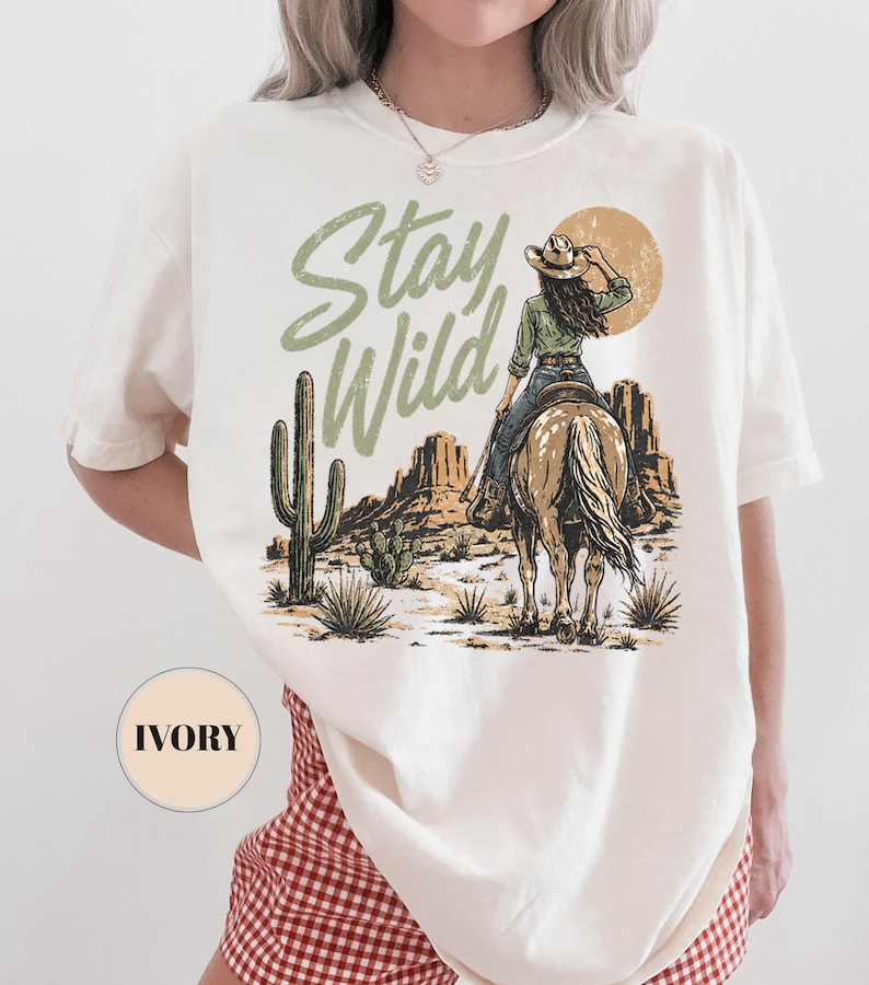 May include: Ivory t-shirt with a desert-themed graphic. It shows a cowgirl on horseback, a cactus, and the words "Stay Wild" in green. The design also includes a sun and mountains.