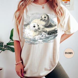 Pode incluir: Camiseta cor marfim com uma ilustra&ccedil;&atilde;o vintage de duas focas numa rocha no oceano. O gr&aacute;fico &eacute; em preto e branco, com linhas detalhadas. A camiseta tem um corte folgado e mangas curtas. A palavra "IVORY" est&aacute; impressa.