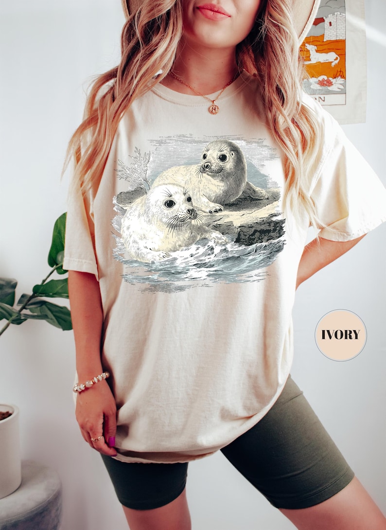 Pode incluir: Camiseta cor marfim com uma ilustra&ccedil;&atilde;o vintage de duas focas numa rocha no oceano. O gr&aacute;fico &eacute; em preto e branco, com linhas detalhadas. A camiseta tem um corte folgado e mangas curtas. A palavra "IVORY" est&aacute; impressa.